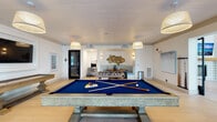 This single-floor amenity space is thoughtfully designed to offer both entertainment and relaxation in a cohesive, coastal-inspired aesthetic. The layout flows between two primary zones: an expansive game room and an open lounge area, connected by wide, light-toned hardwood flooring that runs throughout.

The game room is generously proportioned, anchored by a blue felt billiards table with a substantial pedestal base and complemented by a foosball table and a shuffleboard table. Woven pendant lights hang overhead from a shiplap ceiling, lending warmth and texture to the space. A wall-mounted flat-screen television is framed within a built-in white surround, while a curved banquette seating nook — upholstered in soft blue fabric and adorned with decorative woven wall art — provides an intimate gathering spot. A cue rack and small bistro tables complete the room's functional offerings.

The adjacent lounge area commands attention with dramatic double-height ceilings clad in botanical-patterned wallcovering. Large windows flood the space with natural light, highlighting the neutral furnishings, layered textiles, and a mix of seating that includes sofas, accent chairs, and woven ottomans. A rattan credenza and curated artwork add refined finishing touches throughout.