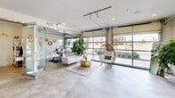 Located in Jenks, Oklahoma, this single-floor community space offers a thoughtfully designed, open-concept layout that maximizes functionality within a compact footprint. The interior is anchored by polished concrete floors that run throughout, lending a clean, industrial aesthetic that pairs well with the bright white walls and modern finishes.

A standout architectural feature is the full-width glass garage door along one wall, which opens the space to abundant natural light and provides clear sightlines to the outdoor patio and surrounding streetscape. This seamless indoor-outdoor connection creates an inviting, airy atmosphere. A glass-paneled partition wall further enhances the sense of openness while defining separate functional zones within the space.

The kitchen is well-appointed with warm wood cabinetry, white quartz countertops, white subway tile backsplash, and floating wood shelves. A bold blue SMEG refrigerator serves as a vibrant focal point, complemented by a dishwasher, microwave, and coffee station. A casual work area with a wooden desk and modern seating is positioned nearby, accented by a decorative "GATHER" sign that reinforces the communal spirit of the space.

The patio extends the usable area outdoors, while the main lounge area features comfortable seating, lush greenery, and a rattan egg chair, creating a welcoming environment ideal for gathering and collaboration.