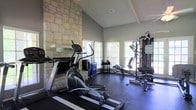 This dedicated fitness space combines rustic charm with functional design, creating an inviting environment for exercise and wellness. The room's most striking architectural feature is the floor-to-ceiling natural stone accent wall, which lends a warm, textural contrast to the smooth grey walls throughout. Soaring vaulted ceilings with decorative beam detailing amplify the sense of openness and airiness within the space. Multiple windows with slatted blinds line the walls, flooding the interior with natural light and offering views of the surrounding outdoors, enhancing the overall workout atmosphere. The flooring is finished with dark, durable rubber gym matting — practical and well-suited for a high-use fitness environment. The space is outfitted with a variety of exercise equipment, including an elliptical cross-trainer, a rowing machine, and a multi-station cable and weight machine, providing a comprehensive range of cardio and strength training options. Additional free weights and accessories are neatly arranged along the perimeter. The combination of architectural elegance and purposeful fitness amenities makes this a standout space that is both visually appealing and thoroughly functional.