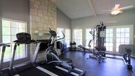 This dedicated fitness space combines rustic charm with functional design, creating an inviting environment for exercise and wellness. The room's most striking architectural feature is the floor-to-ceiling natural stone accent wall, which lends a warm, textural contrast to the smooth grey walls throughout. Soaring vaulted ceilings with decorative beam detailing amplify the sense of openness and airiness within the space. Multiple windows with slatted blinds line the walls, flooding the interior with natural light and offering views of the surrounding outdoors, enhancing the overall workout atmosphere. The flooring is finished with dark, durable rubber gym matting — practical and well-suited for a high-use fitness environment. The space is outfitted with a variety of exercise equipment, including an elliptical cross-trainer, a rowing machine, and a multi-station cable and weight machine, providing a comprehensive range of cardio and strength training options. Additional free weights and accessories are neatly arranged along the perimeter. The combination of architectural elegance and purposeful fitness amenities makes this a standout space that is both visually appealing and thoroughly functional.