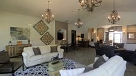 This single-floor space makes an immediate impression with its expansive, open-concept layout and carefully curated design elements. The main living area is anchored by multiple ornate wrought-iron chandeliers that hang from elevated ceilings, casting warm, inviting light throughout the room. Generous seating is arranged around a patterned area rug, creating a comfortable and stylish gathering space. Decorative wall art featuring intricate medallion-style cutouts adds a distinctive artistic touch to the neutral-toned walls. Arched doorways frame the transitions between areas, adding architectural character and a sense of elegance to the overall design. Beyond the living area, a dining section with dark bistro-style tables and chairs provides a defined yet open space for meals or gatherings. Further back, a service or bar area is visible, suggesting versatility in how the space can be used. The flooring throughout is a cool-toned large-format tile that unifies the space and complements the neutral color palette of grays, creams, and warm whites. The combination of classic architectural details, refined furnishings, and carefully chosen décor gives this space a sophisticated yet welcoming ambiance suited for both casual use and more formal occasions.