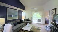 This two-floor residence makes a striking first impression with its soaring vaulted ceilings and dramatic navy blue accent wall in the main living area. The living room is generously proportioned, bathed in natural light that streams through large sliding glass doors dressed with flowing white sheer curtains. These doors open directly onto a private balcony, offering picturesque views of surrounding greenery and creating a seamless connection between the interior and the outdoors. A ceiling fan overhead adds both style and functionality to the space. The living area features plush carpeting underfoot, complemented by a layered area rug that adds warmth and texture to the room. The carefully curated furnishings — including a comfortable sectional sofa, coordinating accent chairs, and a clean-lined coffee table — highlight the space's versatility and livability. The interplay of the crisp white ceiling against the bold accent wall creates a contemporary yet inviting atmosphere. Spanning two floors, the residence offers ample room for comfortable living, with thoughtful design details throughout that balance modern aesthetics with everyday functionality.