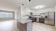 This thoughtfully designed 1-bedroom, 1-bathroom unit offers a clean, modern aesthetic across a single floor. Light-toned hardwood flooring runs throughout, creating a cohesive and bright atmosphere in every room.

The kitchen is a standout feature, showcasing two-tone cabinetry with crisp white upper cabinets and darker lower cabinets accented by gold hardware. A striking marble-look backsplash extends across the full wall, complementing the quartz peninsula countertop. Stainless steel appliances — including a gas range, microwave, dishwasher, and refrigerator — complete the functional yet stylish space. Pendant lighting with gold accents adds a warm, contemporary touch.

The living room is bright and welcoming, with a large window framing an outdoor view and a neutral two-tone wall palette that adds depth to the open space. The bedroom offers a comfortable footprint, with multiple windows that allow natural light to fill the room. A dedicated closet provides ample storage with a full-width hanging rod and built-in shelving.

The bathroom features a modern gray vanity with a quartz countertop, an undermount sink, and matte black fixtures throughout, including the faucet, mirror frame, towel bars, and shower hardware. A tub-shower combination with white tile surround completes the clean, cohesive design.