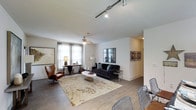 This well-appointed 2-bedroom, 2-bathroom residence offers a thoughtfully designed single-floor layout that maximizes both comfort and functionality. The open-concept living area serves as the heart of the home, featuring rich hardwood-style flooring that flows seamlessly throughout the space. A ceiling fan overhead and track lighting provide flexible illumination, complementing the generous natural light that pours in through large windows dressed with sheer curtains. The living room is generously proportioned, offering ample room for distinct lounging and dining or workspace arrangements. A plush area rug anchors the seating area, creating a warm and inviting atmosphere. The neutral, light-toned walls provide a versatile backdrop that enhances the sense of openness. Wall art adds curated personality to the space, while the transition between rooms is smooth and intuitive. A hallway leads to the private quarters, maintaining a clear separation between the living and sleeping areas. The two bedrooms offer comfortable retreats, as glimpsed through the open doorway revealing a well-styled room with soft furnishings. The overall ambiance is modern and refined, blending contemporary design elements with a welcoming, livable aesthetic throughout.