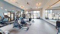 This fitness center in Glendale, Arizona offers a generously sized, single-floor exercise space designed to support a comprehensive workout experience. The main exercise room is bright and open, featuring large floor-to-ceiling windows along one wall that flood the space with natural light and provide views of the surrounding outdoor environment. The flooring transitions between durable rubber gym flooring in the main workout area and carpet in the stretching and cool-down zone, catering to a variety of fitness activities.

The facility is well-stocked with a diverse selection of equipment, including multiple Spirit-brand elliptical trainers and treadmills, a multi-functional cable and strength training rack, a medicine ball storage rack, and a full dumbbell rack with an extensive range of free weights. Ceiling fans and recessed lighting provide comfortable climate control and consistent illumination throughout.

A large wall-mounted mirror with a teal accent wall enhances the sense of depth and space within the exercise room. A separate carpeted flex room with interior-facing glass windows offers a quieter area ideal for stretching, yoga, or functional training. The entry area features glass-panel walls that offer a clear sightline into the gym floor. The overall design is modern and clean, creating an inviting and motivating environment for fitness enthusiasts.