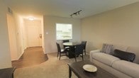 This single-floor space presents an open, functional layout where the living, dining, and kitchen areas flow seamlessly together. The living area is carpeted in a neutral tone and accommodates a sofa and coffee table, creating a comfortable lounge space. A track lighting fixture overhead provides focused illumination, complementing the warm, neutral-toned walls throughout.

Adjacent to the living area, the dining space features a round dark-wood table paired with matching dark chairs, positioned directly in front of a pass-through window that opens into the kitchen. This architectural detail enhances the sense of connectivity between spaces while allowing natural light from the kitchen to filter through.

The kitchen, visible through the pass-through, features white cabinetry and light countertops, offering a clean and practical workspace. A hallway extends from the main living area toward what appears to be additional rooms and the entry door, where a fire extinguisher is mounted — a practical safety feature. The combination of carpet and hard flooring in the hallway adds subtle visual contrast throughout the space.