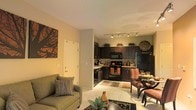 This single-floor residence offers an open-concept layout that seamlessly connects the living, dining, and kitchen areas into one cohesive and functional space. The living area features light tile flooring and is furnished with a sage green sofa accented by decorative pillows, complemented by warm amber-toned walls and bold artwork that add character and depth to the room.

The kitchen is positioned toward the rear of the open layout and is equipped with dark espresso-toned cabinetry that extends to the upper walls, providing generous storage space. Black appliances, including a refrigerator, range, and built-in microwave, offer a sleek, uniform aesthetic. Light-toned countertops contrast nicely with the dark cabinetry, while decorative plants atop the upper cabinets introduce a natural element. Overhead, a track lighting system illuminates the kitchen efficiently.

Adjacent to the kitchen, the dining area features a dark wood table with upholstered chairs, creating an intimate setting for meals. A patterned area rug anchors the dining space, adding texture and warmth. The overall ambiance of the space is cozy and inviting, with a consistent warm color palette tying each area together harmoniously.