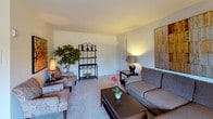 This single-floor residence offers a thoughtfully arranged layout that flows comfortably from room to room. The living room serves as the heart of the home, featuring neutral-toned carpeting and soft ambient lighting that create a warm, inviting atmosphere. The space accommodates a full seating arrangement, complemented by decorative accents including a metal étagère and indoor greenery, lending the room a lived-in, comfortable character.

Adjacent to the living area, the dining room is centered around a classic round wood table with elegantly carved chairs, set beneath a window dressed with horizontal blinds and draping curtains. Natural light filters in, brightening the space throughout the day.

The kitchen is efficiently designed with dark wood cabinetry, light wood-toned countertops, and white tile flooring. It provides ample storage and counter space, making it a functional and organized cooking environment.

The two bedrooms are carpeted and similarly appointed, each offering closet storage and windows that allow natural light. The primary bedroom is notably spacious, featuring room for a full workspace alongside the sleeping area.

The bathroom maintains a clean, classic look with tile flooring, a tub-shower combination, and white vanity cabinetry. Overall, this residence presents a cohesive, functional, and comfortably styled living environment throughout its single level.