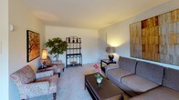 This single-floor residence offers a thoughtfully arranged layout that flows comfortably from room to room. The living room serves as the heart of the home, featuring neutral-toned carpeting and soft ambient lighting that create a warm, inviting atmosphere. The space accommodates a full seating arrangement, complemented by decorative accents including a metal étagère and indoor greenery, lending the room a lived-in, comfortable character.

Adjacent to the living area, the dining room is centered around a classic round wood table with elegantly carved chairs, set beneath a window dressed with horizontal blinds and draping curtains. Natural light filters in, brightening the space throughout the day.

The kitchen is efficiently designed with dark wood cabinetry, light wood-toned countertops, and white tile flooring. It provides ample storage and counter space, making it a functional and organized cooking environment.

The two bedrooms are carpeted and similarly appointed, each offering closet storage and windows that allow natural light. The primary bedroom is notably spacious, featuring room for a full workspace alongside the sleeping area.

The bathroom maintains a clean, classic look with tile flooring, a tub-shower combination, and white vanity cabinetry. Overall, this residence presents a cohesive, functional, and comfortably styled living environment throughout its single level.
