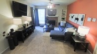 This single-floor residence centers around a welcoming and well-appointed living room that blends comfort with character. The room is anchored by a classic brick fireplace, which serves as a natural focal point and adds a warm, traditional charm to the space. Flanking the fireplace are built-in shelving units, offering both display space and functional storage. A bold terracotta accent wall adds a pop of warmth and color, contrasting beautifully with the neutral tones found throughout the rest of the room. Soft, plush carpeting underfoot contributes to the cozy atmosphere, while a large window dressed with floor-length curtains invites generous natural light into the space. The living area is furnished with a spacious sectional sofa and a chaise, arranged to encourage relaxed conversation or leisurely lounging. A ceiling fan with an integrated light fixture provides both air circulation and ambient lighting. A wall-mounted flat-screen television and a media console round out the entertainment setup, while thoughtful décor — including greenery, accent pillows, and framed artwork — give the space a curated, lived-in feel.