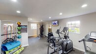 This single-floor fitness center, identified as The Lenox, offers a comprehensive workout environment designed to support a wide range of exercise routines. The space is thoughtfully organized into distinct functional zones, allowing users to move seamlessly between cardio, strength, and flexibility training.

The cardio area is well-stocked with multiple elliptical trainers and a treadmill, positioned to take advantage of natural light streaming in through several windows. A wall-mounted flat-screen television provides entertainment during workouts. Recessed lighting throughout ensures the space remains bright and inviting at all times.

The strength training section features a Hoist multi-functional cable machine paired with an adjustable bench, ideal for a full upper and lower body workout. A dedicated free weight station includes a rack of dumbbells in varying sizes, catering to users of all fitness levels. An informational workout poster is displayed nearby for reference.

A separate flexibility and functional fitness area rounds out the space, equipped with an organized rack holding stability balls, resistance bands, foam rollers, and yoga mats. A wall-mounted mirror allows users to monitor their form. The space features durable carpet flooring, neutral-toned walls, and a clean, functional layout that creates a focused and motivating atmosphere.
