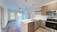 This single-floor residence at 632 SW 150th St in Burien, WA presents a thoughtfully designed open-concept layout finished with contemporary materials throughout. Light wood-tone flooring flows consistently across all rooms, creating a cohesive and airy atmosphere.

The kitchen serves as a central focal point, featuring a substantial island with a striking white quartz countertop accented by subtle veining. Warm wood upper cabinetry contrasts with sleek grey lower cabinets, while stainless steel appliances—including a refrigerator, range, and microwave—add a polished finish. Globe pendant lights with brass fixtures hang above the island, complementing recessed ceiling lighting. Open floating shelves provide additional storage and display space.

The living area extends naturally from the kitchen and opens directly onto a private balcony through sliding glass doors, offering views of surrounding greenery. A ceiling fan overhead adds comfort to the space.

Bedrooms are bright and well-proportioned, each featuring large windows with blinds that frame outdoor views. The hallway connecting the rooms is clean and well-lit with recessed lighting.

The bathroom includes a modern floating vanity with white countertops, a large mirror, and a walk-in shower enclosed by sliding glass doors with chrome hardware. The overall design is clean, minimal, and move-in ready.