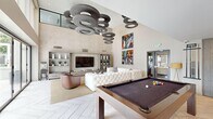 This contemporary single-floor amenity space seamlessly integrates a living room, dining area, and kitchen into one sophisticated, open-plan environment. The living area commands attention with dramatic soaring ceilings adorned by a striking sculptural ring chandelier, creating a bold architectural focal point overhead. A plush tufted white sectional sofa anchors the lounge area alongside a leather accent chair, a herringbone-patterned tile floor, and vibrant abstract artwork displayed on tall neutral walls. A built-in media unit with open shelving adds both function and style. A full-size pool table at the center of the room enhances the space's social and recreational appeal. Expansive floor-to-ceiling sliding glass doors flood the interior with natural light and open directly onto an outdoor patio, offering seamless indoor-outdoor flow and courtyard views. A dedicated dining nook features a rich blue velvet banquette, a round glass table, and curated artwork, providing an intimate setting within the larger open layout. The kitchen is appointed with dark cabinetry, a gleaming white quartz waterfall island with seating for multiple guests, pendant lighting, stainless steel appliances, and a dazzling mosaic tile backsplash. Together, these spaces deliver a refined, design-forward environment ideal for gathering and entertaining.