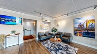 Located at 5 Grandview Ave in Pittsburgh's Mount Washington neighborhood, this well-appointed single-floor space combines polished design with practical functionality. The entrance greets visitors through a striking glass-and-steel vestibule framed by stone accents and flanked by metal railings, with a geometric chandelier overhead that sets an elegant tone from the moment of arrival. The entry foyer transitions seamlessly into a spacious lobby featuring large-format tile flooring, warm wall sconces, and a Pittsburgh skyline artwork that celebrates the local character of the space. Crown molding and track lighting add refined architectural detail throughout. The waiting area is thoughtfully furnished with tufted leather chairs arranged around a modern coffee table atop a layered area rug, all set on rich hardwood flooring. Vibrant, large-scale artwork — including a colorful city map print and a vivid photograph of Pittsburgh's iconic Roberto Clemente Bridge — gives the space an inviting, curated ambiance. A reception desk area provides a functional check-in point, while a dedicated fitness room rounds out the amenities, equipped with a treadmill, medicine ball rack, wall-mounted television, and a mini-split HVAC unit. Glass block windows provide natural light while maintaining privacy in the fitness space. Overall, the space strikes a balance between professional polish and welcoming comfort.