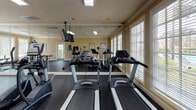 This fitness center at 9001 SR-21 S in Port Wentworth, Georgia, is a well-appointed single-story space designed to support a comprehensive workout experience. The room features warm beige walls accented by crisp crown molding, dark rubber flooring, and a bright, open layout illuminated by a combination of recessed lighting and large fluorescent ceiling fixtures.

Natural light flows generously into the space through multiple sets of large windows fitted with white blinds, offering views of the surrounding greenery. A full-length wall mirror on one side of the room visually expands the space and allows users to monitor their form during exercise.

The cardio section is well-stocked with multiple True-brand treadmills and elliptical trainers, positioned to take advantage of the natural light from the windows. A wall-mounted flat-screen television is suspended from the ceiling for entertainment during workouts. The strength training area features a multi-station cable machine, a flat bench, and additional free weight equipment, providing a well-rounded selection for resistance training.

Informational fitness posters are mounted on the walls, and a scale is available near the entrance. The space is monitored by video surveillance and includes clearly marked emergency exits, ensuring a safe and functional environment for all users.