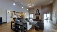 This single-floor residence showcases an expansive open-concept design that effortlessly connects the living, dining, and kitchen areas into one cohesive and inviting space. The great room commands immediate attention with its dramatically soaring ceilings, punctuated by clerestory windows that flood the interior with abundant natural light. A grand chandelier with warm Edison-style bulbs serves as a striking focal point overhead, complementing the refined yet comfortable aesthetic throughout.

The living area is anchored by a floor-to-ceiling natural stone fireplace, lending a rustic yet sophisticated character to the room. Tufted gray sofas and elegant white accent chairs are arranged around a glass-top coffee table, creating an ideal setting for both relaxed gatherings and more formal entertaining.

Transitioning seamlessly into the kitchen, the space features professional-grade stainless steel appliances, a substantial island with seating, a statement range hood, and a built-in wine rack with distinctive stone detailing. Warm hardwood flooring flows throughout, tying together the various areas with continuity and warmth.

Natural light pours in through large windows and glass doors at the rear, offering views to the outdoors. The overall ambiance is one of understated luxury — warm, spacious, and thoughtfully designed for modern living.