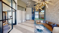 This single-floor communal space, identified as the Foundry at Westover House, delivers a striking blend of industrial and contemporary design throughout its expansive, open layout. The entrance is defined by floor-to-ceiling glass walls and dark-framed double doors that flood the interior with abundant natural light, framing outdoor views of the surrounding streetscape.

The soaring ceilings serve as a dramatic architectural feature, anchored by a large, sculptural sputnik-style chandelier with gold radiating arms and warm Edison-style bulbs. A full-height exposed brick wall provides rich texture and an industrial character, contrasting beautifully with smooth painted surfaces in muted gray and bold cobalt blue tones. Structural columns clad in blue add further visual depth throughout the space.

Light-toned large-format tile flooring extends across the entire layout, creating a seamless, cohesive flow between zones. A welcoming lounge area features a cognac leather sofa and a modern rounded coffee table atop a patterned area rug. Adjacent seating nooks with navy blue upholstered booth seating offer semi-private areas for individual use. A separate conference room with expansive windows provides a functional workspace. Curated artwork and lush greenery complete the polished, contemporary atmosphere.