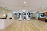 Located at 2855 W Commercial Blvd in Fort Lauderdale, Florida, this single-floor commercial space offers a generously sized, thoughtfully designed interior that blends contemporary aesthetics with practical functionality. The entry foyer welcomes visitors through expansive floor-to-ceiling glass doors that frame views of the surrounding landscaping, flooding the space with natural light and creating a bright, airy first impression. Wide-plank wood-look flooring flows seamlessly throughout, unifying the various zones of the interior.

At the heart of the space is an elegant reception desk with a ribbed white paneled front, flanked by decorative wall-mounted art and sconce lighting. Beyond the reception area, two distinct lounge zones are defined by bold, large-scale abstract murals in navy blue, tan, and cream — a striking design feature that serves as a visual anchor for the space. Curved cream-upholstered chairs, modular seating, and organic accent tables complete these inviting gathering areas. A dramatic multi-ring chandelier overhead adds a sculptural focal point.

A dedicated café-style kitchen area features rich, warm-toned cabinetry, white quartz countertops, a tiled backsplash, stainless steel appliances, pendant lighting, and a commercial refrigerated display case. A long corridor extends toward additional interior rooms, lined with soft ambient lighting that guides visitors deeper into the facility.