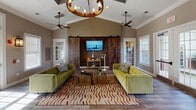 This single-story space combines modern farmhouse aesthetics with functional design across several distinct areas. The main lounge serves as the centerpiece, showcasing soaring vaulted ceilings accented by crown molding and dual ceiling fans. A striking pair of rustic sliding barn doors frame a mounted flat-screen TV, while two plush olive-green velvet sofas face each other atop a bold chevron-patterned area rug. A statement wagon-wheel chandelier anchors the room, and large French doors with transom windows flood the space with natural light, offering views of the surrounding outdoor area.

Adjacent to the lounge, a casual dining nook features a round wood-top table with upholstered chairs, positioned near additional French doors that provide a seamless connection to the exterior. The warm hardwood-style flooring flows consistently throughout the space.

A private office area features vaulted ceilings, interior pass-through windows, and a substantial dark wood desk, creating a professional yet welcoming workspace. The neutral taupe wall palette, white trim, and carefully curated décor unify the space, lending it a polished, contemporary character throughout.