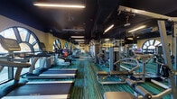 Located at 2500 Longview St in Austin, TX, this fitness center offers a comprehensive workout environment across a single, well-organized floor. The space is defined by its dark ceiling with linear LED lighting, warm yellow walls, and vibrant teal-toned multi-stripe carpet flooring, creating an energetic yet focused atmosphere.

The facility is generously outfitted with a wide array of cardio equipment, including multiple TRUE-brand treadmills and stationary bikes, all positioned to take advantage of the room's striking arched windows. These large, architecturally distinctive windows flood the space with natural light and provide open views of the surrounding outdoor greenery.

Along the mirrored walls, a full selection of Technogym strength machines and cable systems are arranged to allow for efficient, circuit-style training. A dedicated free weights section features a complete dumbbell rack alongside exercise balls and yoga mats, supporting stretching and functional fitness routines. Wall-mounted televisions are positioned throughout for entertainment during workouts.

A built-in shelving unit and cabinetry offer convenient storage, while a compact refrigeration unit supports hydration needs. The space also includes access to an adjacent restroom facility. Overall, the fitness center strikes a balance between functionality and aesthetic appeal, making it a well-rounded destination for a full-body workout.