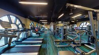 Located at 2500 Longview St in Austin, TX, this fitness center offers a comprehensive workout environment across a single, well-organized floor. The space is defined by its dark ceiling with linear LED lighting, warm yellow walls, and vibrant teal-toned multi-stripe carpet flooring, creating an energetic yet focused atmosphere.

The facility is generously outfitted with a wide array of cardio equipment, including multiple TRUE-brand treadmills and stationary bikes, all positioned to take advantage of the room's striking arched windows. These large, architecturally distinctive windows flood the space with natural light and provide open views of the surrounding outdoor greenery.

Along the mirrored walls, a full selection of Technogym strength machines and cable systems are arranged to allow for efficient, circuit-style training. A dedicated free weights section features a complete dumbbell rack alongside exercise balls and yoga mats, supporting stretching and functional fitness routines. Wall-mounted televisions are positioned throughout for entertainment during workouts.

A built-in shelving unit and cabinetry offer convenient storage, while a compact refrigeration unit supports hydration needs. The space also includes access to an adjacent restroom facility. Overall, the fitness center strikes a balance between functionality and aesthetic appeal, making it a well-rounded destination for a full-body workout.