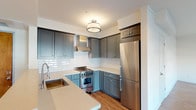 This single-floor unit offers a thoughtfully designed layout with a cohesive aesthetic carried throughout. The kitchen stands as a highlight of the space, showcasing two-toned gray shaker cabinetry — deeper slate uppers paired with softer blue-gray lowers — complemented by white subway tile backsplash, light quartz countertops, and stainless steel appliances including a gas range and refrigerator. A undermount sink and pendant-style flush mount lighting complete the polished look.

The open living area extends from the kitchen, featuring warm wood-look flooring that runs continuously through the space, white walls, and large windows fitted with roller shades that invite natural light and street views. The layout flows naturally between the living and sleeping areas, with the bedrooms offering a clean, neutral palette, ample space, and decorative flush mount fixtures overhead. One bedroom features a walk-in closet and a sizable window overlooking the surrounding neighborhood.

The bathroom is finished with white subway tile surrounding a soaking tub and shower combination, accented by a mosaic border detail, while charcoal hexagonal floor tile adds visual contrast. The overall ambiance is modern yet warm, with consistent finishes creating a unified and comfortable living environment.