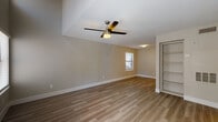 This two-floor space offers a clean, modern aesthetic defined by warm wood-look flooring that runs consistently throughout. The main living area is generously proportioned, featuring a vaulted ceiling detail that adds architectural interest and a sense of openness. A ceiling fan with integrated lighting serves as a functional focal point overhead, while natural light filters in through blinded windows. Built-in open shelving is positioned along one wall, providing convenient storage and display space. The galley-style kitchen is efficiently designed with white shaker cabinetry extending to the ceiling, dark countertops, a classic subway tile backsplash, and a full suite of stainless steel appliances including a refrigerator, dishwasher, electric range, and microwave. Track lighting illuminates the workspace effectively. A bedroom on the upper level benefits from its own ceiling fan, multiple windows with natural views of surrounding trees, and an open half-wall detail that enhances the sense of vertical space. The bathroom includes a full tub-shower combination with white tile surround and chrome fixtures. Neutral wall tones and white trim throughout contribute to a cohesive, understated ambiance.