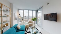 This thoughtfully designed high-rise unit offers a compelling blend of contemporary style and urban sophistication across a single floor. The living area is the centerpiece of the space, anchored by dramatic floor-to-ceiling corner windows that frame panoramic city skyline views and flood the room with natural light. The open layout accommodates a vibrant teal sofa, a curved accent chair, and a glass coffee table atop a bold chevron rug, creating a dynamic and inviting atmosphere. The kitchen is sleek and functional, featuring light wood cabinetry that extends to the ceiling, stone countertops, a generous breakfast bar with seating, light hardwood-style flooring, and recessed lighting that keeps the space bright and airy. The bedroom is a standout, distinguished by a rich emerald green accent wall adorned with curated artwork and flanked by brass swing-arm sconces. A large sliding glass door connects the bedroom directly to a private balcony with city views, while a built-in shelving unit and sliding closet provide ample storage. The bathroom is clean and modern, finished with terrazzo-style flooring, a glass-enclosed walk-in shower, a wood-framed vanity with open shelving, and crisp white walls that lend a spa-like quality to the space.