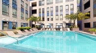The Pool Courtyard amenity complex delivers a well-rounded recreational experience across two primary areas: an outdoor pool courtyard and an adjacent fitness center. The outdoor pool area features a large, pristine swimming pool with crystal-clear turquoise water, surrounded by a generous pool deck lined with cushioned lounge chairs and shaded by multiple patio umbrellas. Lush tropical palms and manicured greenery frame the courtyard, while the multi-story building facade enclosing the space creates a private, resort-like atmosphere bathed in natural sunlight. The fitness center is a spacious, well-lit facility with light wood-toned flooring and floor-to-ceiling mirrors that visually expand the space. It is comprehensively equipped with a full lineup of cardio machines including treadmills, ellipticals, and stationary bikes, as well as a complete dumbbell rack, medicine balls, and multi-station cable and weight machines. Built-in wooden shelving provides organized storage for rolled towels and accessories. A ceiling fan ensures comfortable airflow, and large windows and glass doors offer direct views of the pool courtyard, seamlessly connecting the indoor and outdoor amenity spaces. Recessed lighting throughout keeps the environment bright and energizing.