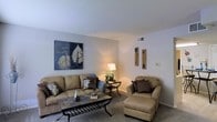 This single-floor space centers around a welcoming living room designed with comfort and functionality in mind. The room features neutral-toned walls that create a clean, open backdrop, complemented by soft carpeted flooring that adds warmth underfoot. A textured ceiling adds subtle architectural interest throughout the space.

The living area is thoughtfully arranged, anchored by a plush tan sofa accented with decorative pillows, paired with a matching armchair and ottoman that invite relaxation. A slate-topped coffee table with a wrought iron base serves as a stylish focal point, offering both visual appeal and practicality. A small accent table with a warm-toned lamp provides ambient lighting, enhancing the room's cozy atmosphere.

Wall décor includes a bold botanical canvas print on one side and a set of vertically arranged wooden arrow accents on the opposite wall, adding personality and visual balance to the space. A ceiling vent indicates a central HVAC system for year-round climate comfort. The overall ambiance is warm, inviting, and well-coordinated, making it a functional and aesthetically pleasing living environment.