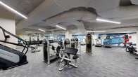 This expansive fitness center offers a comprehensive workout environment spread across a single floor. The space is defined by an industrial-style ceiling with exposed ductwork and structural elements, illuminated throughout by rows of sleek linear LED fixtures that provide consistent, bright lighting across all workout zones.

The facility is organized into distinct functional areas. A dedicated cardio section lines one wall, housing multiple treadmills, elliptical trainers, stationary bikes, and a stair climber. Adjacent to this, a free weights area features a full rack of dumbbells paired with adjustable benches. Strength training is further supported by a selection of cable and resistance machines distributed throughout the floor.

A functional training corner near the entrance is stocked with stability balls, medicine balls, resistance bands, and a multi-purpose cable machine, catering to dynamic and rehabilitative exercises. The entry area features glass doors and a posted fitness center rules board, contributing to a professional atmosphere.

Throughout the space, a vibrant abstract mural in cool blues and warm tones spans the perimeter walls, adding an energizing visual element. The flooring consists of durable patterned carpet tiles, suitable for high-traffic use. The overall layout is open and navigable, with clearly marked floor indicators supporting organized use of the facility.