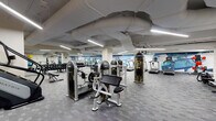 This expansive fitness center offers a comprehensive workout environment spread across a single floor. The space is defined by an industrial-style ceiling with exposed ductwork and structural elements, illuminated throughout by rows of sleek linear LED fixtures that provide consistent, bright lighting across all workout zones.

The facility is organized into distinct functional areas. A dedicated cardio section lines one wall, housing multiple treadmills, elliptical trainers, stationary bikes, and a stair climber. Adjacent to this, a free weights area features a full rack of dumbbells paired with adjustable benches. Strength training is further supported by a selection of cable and resistance machines distributed throughout the floor.

A functional training corner near the entrance is stocked with stability balls, medicine balls, resistance bands, and a multi-purpose cable machine, catering to dynamic and rehabilitative exercises. The entry area features glass doors and a posted fitness center rules board, contributing to a professional atmosphere.

Throughout the space, a vibrant abstract mural in cool blues and warm tones spans the perimeter walls, adding an energizing visual element. The flooring consists of durable patterned carpet tiles, suitable for high-traffic use. The overall layout is open and navigable, with clearly marked floor indicators supporting organized use of the facility.