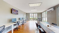 This single-floor community workspace offers a well-appointed and functional environment designed to support a variety of activities. The space is finished with warm hardwood flooring throughout, complemented by neutral gray walls and a clean white ceiling fitted with recessed panel lighting that ensures consistent illumination across the entire area.

Along one wall, a long white workstation desk accommodates multiple computer terminals, each paired with ergonomic rolling chairs, making it ideal for residents or members needing a dedicated work or study setup. A wall-mounted flat-screen television is positioned above the workstation area for shared viewing. A printer station is also available nearby, adding to the practical amenities on offer.

A rustic wood communal table with seating provides an alternative workspace or gathering area, while a bistro-style high-top table and stools are situated in the sunlit corner. This corner is defined by expansive floor-to-ceiling windows on two walls, offering panoramic views of the surrounding landscaped grounds and filling the space with abundant natural light.

Deeper into the space, a pool table adds a recreational dimension, catering to leisure and social interaction. An additional wall-mounted screen and recessed lighting continue in this area. A door provides direct access to an outdoor covered walkway, further enhancing the connection to the exterior environment. Overall, the space balances productivity and recreation in a bright, open setting.