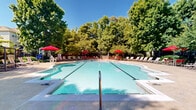 Located at 721 S Cherry Grove Ave in Annapolis, Maryland, this outdoor amenity area centers around a generously sized swimming pool with clear, blue-green water and defined lap lanes. The pool features both shallow and deeper sections, with a gradual depth transition clearly marked along the pool edge, making it suitable for a range of swimmers.

Surrounding the pool is a expansive concrete sun deck lined with striped lounging chairs, offering ample space for relaxation under the open sky. Vibrant red market umbrellas are positioned throughout the deck, providing shaded seating areas with coordinating red chairs and tables — ideal for socializing or unwinding outdoors.

The perimeter of the pool area is enclosed by a black iron fence, ensuring privacy and security while maintaining an open, airy feel. Mature deciduous trees frame the space on all sides, offering natural shade and a lush, park-like atmosphere that softens the hardscape. A brick clubhouse building is visible at one end of the pool, adding architectural character to the setting.

The overall ambiance is bright, welcoming, and well-maintained, reflecting a community-oriented outdoor retreat designed for warm-weather enjoyment. The combination of generous seating, mature greenery, and classic design elements makes this pool area a standout feature of the Cherry Grove Avenue property in Annapolis.