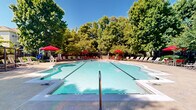Located at 721 S Cherry Grove Ave in Annapolis, Maryland, this outdoor amenity area centers around a generously sized swimming pool with clear, blue-green water and defined lap lanes. The pool features both shallow and deeper sections, with a gradual depth transition clearly marked along the pool edge, making it suitable for a range of swimmers.

Surrounding the pool is a expansive concrete sun deck lined with striped lounging chairs, offering ample space for relaxation under the open sky. Vibrant red market umbrellas are positioned throughout the deck, providing shaded seating areas with coordinating red chairs and tables — ideal for socializing or unwinding outdoors.

The perimeter of the pool area is enclosed by a black iron fence, ensuring privacy and security while maintaining an open, airy feel. Mature deciduous trees frame the space on all sides, offering natural shade and a lush, park-like atmosphere that softens the hardscape. A brick clubhouse building is visible at one end of the pool, adding architectural character to the setting.

The overall ambiance is bright, welcoming, and well-maintained, reflecting a community-oriented outdoor retreat designed for warm-weather enjoyment. The combination of generous seating, mature greenery, and classic design elements makes this pool area a standout feature of the Cherry Grove Avenue property in Annapolis.