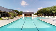 The pool and outdoor lounge area is a thoughtfully designed amenity space that combines resort-style recreation with functional gathering areas. The expansive swimming pool serves as the central feature, offering dedicated lap lanes for fitness swimming alongside a shallow lounge section equipped with sleek, modern in-pool chairs—ideal for relaxation. The pool deck is finished in large-format neutral tile, providing ample space for sunbathing with multiple lounge chairs positioned around the perimeter.

A standout architectural element is the adjacent covered cabana pavilion, constructed with a steeply pitched, natural wood-planked ceiling that lends warmth and character to the outdoor space. This covered lounge features a floor-to-ceiling hexagonal-tile fireplace, a wall-mounted television, and a built-in wet bar with refrigeration units—creating a comfortable gathering environment in any season. Ceiling fans ensure airflow throughout.

A separate outdoor kitchen station includes dual stainless steel grills set into a hexagonal-tiled counter with granite surfaces, designed for community grilling and entertaining. The surrounding landscape features mature shade trees, softening the space and providing natural privacy. The metal standing-seam roof of the poolhouse ties the architectural elements together, creating a cohesive, upscale outdoor environment.