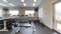 This fitness center is thoughtfully designed to accommodate a wide range of workout routines within a bright, functional space. The interior features neutral beige walls accented by white crown molding, durable rubber flooring, and recessed fluorescent lighting that ensures the space is consistently well-lit. Large windows fitted with white horizontal blinds allow natural light to filter in while maintaining privacy, and views of the surrounding outdoors add a pleasant backdrop to workouts.

The cardio zone is equipped with multiple treadmills, elliptical trainers, a Precor elliptical, and a Bodycraft rowing machine, providing diverse options for aerobic exercise. A wall-mounted television is positioned for easy viewing during cardio sessions. Full-length mirrors line the walls, visually expanding the space and allowing users to monitor their form throughout their workouts.

A dedicated strength training area houses a multi-function cable machine, an adjustable weight bench, a rack of dumbbells in varying weights, and a medicine ball storage tower with multiple balls. Sanitation supplies are conveniently available near the entrance, reinforcing a clean and hygienic environment. The fitness center is accessed through a glass-paneled door, contributing to an open and welcoming atmosphere.