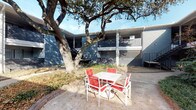 Located at 2770–2798 Teagu in Houston, Texas, this multi-unit residential complex showcases a classic mid-century architectural style characterized by dark blue-grey brick exteriors and a two-story wraparound building configuration. The structure frames a series of interior courtyards that serve as the heart of the community, creating a semi-private outdoor environment sheltered from the surrounding streetscape.

The courtyards are defined by curved brick-edged landscaping beds filled with low shrubs, ornamental grasses, and slender trees that provide seasonal greenery. Mature live oak trees anchor the outdoor spaces, their broad canopies offering natural shade over the concrete patio areas below. A white outdoor dining table with red slatted chairs is positioned beneath one of the oaks, providing a functional and shaded gathering spot for residents.

Concrete walkways wind through the landscaped grounds, connecting building entrances and stairwells across the property. Black metal staircases provide access to the upper-level corridors, which feature covered walkways running the length of each building wing. The overall layout encourages an open-air, community-oriented atmosphere. The combination of established vegetation, brick detailing, and the inward-facing courtyard design gives this Houston property a cohesive and well-maintained residential character.