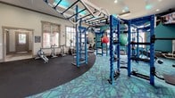 This fitness center is a comprehensively outfitted single-floor facility designed to support a wide range of workout styles and fitness goals. The space is visually striking, anchored by a bold color palette of deep blues and teals that carries through the equipment, accent walls, and patterned carpet flooring. A central functional training rig in vivid blue dominates the main workout area, featuring monkey bars, gymnastic rings, hanging punching bags, medicine balls, and kettlebells — all suspended or mounted within a ceiling-connected frame with teal LED accent lighting above.

Surrounding the central rig, the cardio zone offers treadmills, elliptical trainers, and stationary bikes positioned near large windows that invite natural light and outdoor views. A wall-mounted flat-screen television provides entertainment during cardio sessions. A dedicated dumbbell rack lines one wall alongside additional strength training equipment.

One of the more distinctive features is a rock climbing wall labeled "Freedom Climber," fitted with multicolored hand holds and set against a teal accent wall. Adjacent to it is a water station with a teal penny-tile accent wall, pendant lighting, and access to additional rooms. The facility's high tray ceilings, recessed lighting, and cohesive design create an energizing, modern atmosphere throughout.