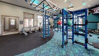 This fitness center is a comprehensively outfitted single-floor facility designed to support a wide range of workout styles and fitness goals. The space is visually striking, anchored by a bold color palette of deep blues and teals that carries through the equipment, accent walls, and patterned carpet flooring. A central functional training rig in vivid blue dominates the main workout area, featuring monkey bars, gymnastic rings, hanging punching bags, medicine balls, and kettlebells — all suspended or mounted within a ceiling-connected frame with teal LED accent lighting above.

Surrounding the central rig, the cardio zone offers treadmills, elliptical trainers, and stationary bikes positioned near large windows that invite natural light and outdoor views. A wall-mounted flat-screen television provides entertainment during cardio sessions. A dedicated dumbbell rack lines one wall alongside additional strength training equipment.

One of the more distinctive features is a rock climbing wall labeled "Freedom Climber," fitted with multicolored hand holds and set against a teal accent wall. Adjacent to it is a water station with a teal penny-tile accent wall, pendant lighting, and access to additional rooms. The facility's high tray ceilings, recessed lighting, and cohesive design create an energizing, modern atmosphere throughout.
