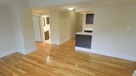 This single-floor space offers a clean, functional layout highlighted by polished hardwood flooring that runs throughout, reflecting light and adding warmth to every room. The open living area is finished in a soft neutral tone, creating a calm and inviting atmosphere that feels both spacious and easy to personalize. A kitchen peninsula with dark cabinetry and a white countertop serves as a natural divider between the living and kitchen areas, providing additional prep space and casual seating options. The dark-toned cabinetry continues into the kitchen, offering a striking contrast against the lighter walls and countertops. A hallway visible from the main living area leads to additional rooms, including a bathroom that features coordinating dark cabinetry and a glimpse of white fixtures, maintaining a consistent design language throughout. Flush-mount ceiling lighting provides practical illumination, while the white baseboards and trim offer a crisp, finished look. The overall aesthetic is modern yet understated, making the space highly versatile for a variety of uses. The thoughtful combination of materials and finishes gives this single-floor layout a cohesive, move-in-ready appeal.