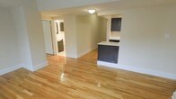 This single-floor space offers a clean, functional layout highlighted by polished hardwood flooring that runs throughout, reflecting light and adding warmth to every room. The open living area is finished in a soft neutral tone, creating a calm and inviting atmosphere that feels both spacious and easy to personalize. A kitchen peninsula with dark cabinetry and a white countertop serves as a natural divider between the living and kitchen areas, providing additional prep space and casual seating options. The dark-toned cabinetry continues into the kitchen, offering a striking contrast against the lighter walls and countertops. A hallway visible from the main living area leads to additional rooms, including a bathroom that features coordinating dark cabinetry and a glimpse of white fixtures, maintaining a consistent design language throughout. Flush-mount ceiling lighting provides practical illumination, while the white baseboards and trim offer a crisp, finished look. The overall aesthetic is modern yet understated, making the space highly versatile for a variety of uses. The thoughtful combination of materials and finishes gives this single-floor layout a cohesive, move-in-ready appeal.