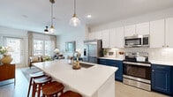 This thoughtfully designed single-floor residence offers an efficient yet stylish layout across a comfortable footprint. The open-concept kitchen and living area serves as the heart of the home, where a large central island with a white quartz countertop and an integrated sink provides both workspace and casual seating. The kitchen features striking two-tone cabinetry — crisp white upper cabinets paired with bold navy blue lower cabinets — complemented by a mosaic tile backsplash and a full suite of stainless steel appliances, including a French door refrigerator, range, microwave, and dishwasher. Light hardwood flooring flows throughout, unifying the space. The living room is bathed in natural light from large windows and a glass door leading to a private balcony, enhanced by a ceiling fan overhead. The bedroom offers a serene retreat with ample natural light from its own set of windows and balcony access, a ceiling fan, and direct access to a dedicated closet. The bathroom features navy blue vanity cabinetry consistent with the home's design theme. Clean white walls and warm wood tones throughout create a cohesive, modern aesthetic with an inviting, residential character.
