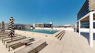 Located in New Orleans, this rooftop pool deck offers a well-appointed outdoor retreat with a modern, urban aesthetic. The centerpiece is a generously sized rectangular lap pool finished in light-toned tile, flanked by wide, tiered pool surround steps and lined with lounge chairs and bold black-and-white striped umbrellas. The clean, neutral palette of the hardscape creates a bright, open atmosphere that maximizes the abundant Louisiana sunlight.

Adjacent to the pool, a pergola-covered lounge and dining area provides welcome shade. The slatted wood overhead structure filters light elegantly, casting geometric shadows across the large-format tile flooring below. Green metal dining chairs and round tables furnish the space, offering functional seating for outdoor gatherings. Floor-to-ceiling glazed panels enclose an interior room accessible from this shaded zone, adding versatility to the space.

Raised planters filled with vibrant flowering plants and greenery are distributed throughout the deck, softening the contemporary architecture. The surrounding New Orleans skyline, including recognizable high-rise towers and the Caesars Superdome, forms a compelling backdrop. The overall ambiance is clean, modern, and inviting, designed for both relaxation and social gatherings in an elevated urban setting.