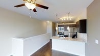 This single-floor space showcases an open-concept design that seamlessly connects the living area to the kitchen, creating a cohesive and functional layout. Dark hardwood floors run throughout, providing a rich contrast against the neutral, light-toned walls and white trim detailing.

The kitchen is a standout feature, equipped with stainless steel appliances including a gas range, built-in microwave, and refrigerator. Dark espresso cabinetry pairs elegantly with a light granite countertop that extends into a peninsula, offering additional prep space and a casual seating area. A classic subway tile backsplash adds texture, while two pendant lights hang above the peninsula, complementing the track lighting overhead that brightens the cooking space.

The living area is generously proportioned, with a ceiling fan providing both comfort and a subtle design element. A second ceiling fan is visible deeper in the space, suggesting ample square footage. White wainscoting-style half-walls frame the transition between areas, adding architectural interest. The overall ambiance is modern yet warm, with layered lighting and quality finishes contributing to a polished, move-in-ready feel.