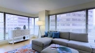 This single-floor residence offers a well-appointed living space defined by clean lines and a contemporary design sensibility. The living area is a standout feature, bathed in abundant natural light streaming through large, floor-to-adjacent-ceiling windows dressed with sleek horizontal blinds. The windows frame views of the surrounding urban landscape, creating a seamless connection between the interior and the city beyond.

The living room is thoughtfully arranged around a generous L-shaped sectional sofa in a soft grey upholstery, accented with coordinating blue throw pillows that add a subtle pop of coastal-inspired color. A matching ottoman doubles as a coffee table, contributing to the room's relaxed yet polished feel. A vibrant area rug featuring a coral and ocean motif anchors the seating arrangement and reinforces the coastal theme.

A white dresser positioned near the windows provides functional storage while keeping the space feeling airy and uncluttered. A modern tripod floor lamp adds a stylish lighting element to the corner of the room. Light-toned flooring runs throughout, enhancing the bright and open atmosphere. The neutral wall palette in soft white tones complements the overall decor, making the space feel welcoming and versatile.