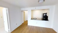 This single-floor residence is designed with a straightforward, functional layout that maximizes the sense of openness throughout its living areas. The main living space features warm hardwood-style flooring that extends seamlessly through the room, complemented by crisp white walls and a textured ceiling with flush-mount lighting overhead.

A defining architectural feature is the pass-through kitchen cutout, which connects the kitchen to the living area. The kitchen is equipped with a stainless steel refrigerator and a dark countertop with a built-in sink, visible through the opening. This design choice creates a sense of connectivity between the two spaces while maintaining a degree of separation.

A folding door off the living area provides access to a hallway leading further into the unit, hinting at additional rooms beyond. The white trim and baseboards throughout lend a clean, cohesive finish to the interior. The overall ambiance is bright and neutral, offering a blank canvas that is well-suited for a variety of interior styles and furnishings. The layout prioritizes practicality and ease of movement throughout the space.