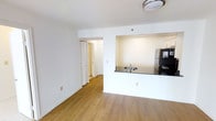 This single-floor residence is designed with a straightforward, functional layout that maximizes the sense of openness throughout its living areas. The main living space features warm hardwood-style flooring that extends seamlessly through the room, complemented by crisp white walls and a textured ceiling with flush-mount lighting overhead.

A defining architectural feature is the pass-through kitchen cutout, which connects the kitchen to the living area. The kitchen is equipped with a stainless steel refrigerator and a dark countertop with a built-in sink, visible through the opening. This design choice creates a sense of connectivity between the two spaces while maintaining a degree of separation.

A folding door off the living area provides access to a hallway leading further into the unit, hinting at additional rooms beyond. The white trim and baseboards throughout lend a clean, cohesive finish to the interior. The overall ambiance is bright and neutral, offering a blank canvas that is well-suited for a variety of interior styles and furnishings. The layout prioritizes practicality and ease of movement throughout the space.