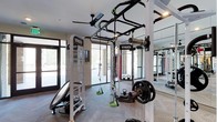 Located at 3100 Women's Club Dr in Raleigh, NC, this single-floor fitness center is a well-appointed gym space designed to support a full range of training disciplines. The facility is anchored by a professional TKO functional training rig, complete with pull-up bars, suspension trainers, resistance bands, and cable attachments — offering a versatile setup for strength, cardio, and bodyweight training. Weight plates, kettlebells, and additional free weights are readily accessible throughout the space. A full-length mirror spans one wall, providing visual feedback during workouts and enhancing the sense of openness in the room. Overhead, a ceiling fan ensures comfortable airflow during intense sessions. The interior is finished with a herringbone-patterned floor and neutral-toned walls that contribute to a clean, modern aesthetic. Large floor-to-ceiling glass doors and windows flood the space with natural light while offering views of the surrounding outdoor environment. The bright, airy atmosphere combined with high-quality equipment creates a motivating and professional training environment suitable for a wide variety of fitness routines.