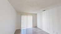 This two-bedroom, one-bathroom residence in San Antonio, Texas offers a well-organized single-floor layout designed for comfortable everyday living. The living room is bright and airy, featuring light wood-style flooring and a large sliding glass door dressed with vertical blinds that invite natural light and provide access to an outdoor area. The space flows seamlessly into a galley-style kitchen, which is outfitted with white shaker cabinetry, black hardware, a single-basin sink, and black and stainless steel appliances, all set over matching light wood-style flooring. Both bedrooms are carpeted and offer built-in closets for ample storage. The primary bedroom is generously sized, complete with a ceiling fan, a window with blinds, and direct access to a vanity area featuring a wide mirror, gray cabinetry, and a well-lit makeup counter. The shared full bathroom includes a tub-shower combination and white overhead cabinetry with brushed nickel hardware. Throughout the home, textured ceilings and crisp white walls contribute to a clean, neutral aesthetic. The second bedroom also includes a standard closet and its own window, adding natural light to the space.